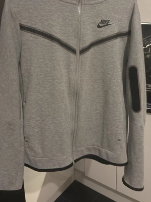 Grå Nike hoodie med dragkedja - Snygg grå hoodie från Nike med hel dragkedja framtill och svarta detaljer. Har en huva och en praktisk ficka med dragkedja på ärmen. 