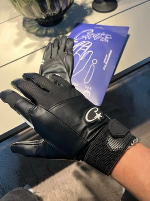 Svarta skinnhandskar från Corteiz - Snygga svarta handskar från Corteiz med vit logga och stjärndetalj. Handskarna har justerbar kardborre vid handleden och är tillverkade i skinn och syntetmaterial för både stil och funktion. Perfekta för dig som vill ha en clean look vid motorcykeln, skidorna eller snowboarden. Låda medföljer och pris kan diskuteras