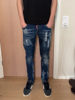 Dsquared2 slim jeans - Säljer ett par blå skinny jeans med slitningar och färgstänk för en trendig look. Jeansen har fem fickor, låg midja och en röd detalj vid gylfen. Perfekta för dig som gillar streetwear och vill sticka ut lite extra.