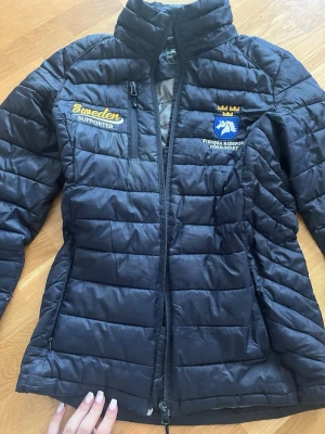 Svart quiltad pufferjacka Sweden Supporter - Ena jackfickan är trasig Svart quiltad pufferjacka med bro sc h Sweden Supporter och Svenska Ridsport solid förbundet på bröstet. Jackan har dragkedja framtill, hög krage och en bröstficka med dragkedja. Perfekt för dig som vill visa ditt stöd för svensk r pen.