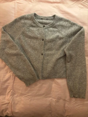 Grå stickad kofta med knappar - Mysig grå stickad kofta med rund halsringning och tre knappar framtill. Koftan har långa ärmar och ribbade muddar vid ärmslut och nederkant. Perfekt för lager på lager och enkel att matcha till olika outfits.