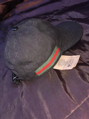 Svart Gucci keps med GG-mönster - Svart keps från Gucci med diskret GG-logomönster över hela tyget. Klassisk böjd skärm och justerbar rem baktill. Sidan pryds av den ikoniska gröna och röda randen. Tillverkad i canvasmaterial som ger en lyxig känsla.