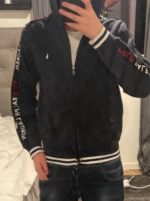 Svart zip hoddie från Ralph Lauren - Säljer en mörkblå hoodie från Ralph Lauren med dragkedja framtill och vit logga på bröstet. Huva. Med  vita detaljer på muddar och nederkant. Text i vitt och rött längs ärmarna ger en sportig vibe. Perfekt för dig som gillar streetwear. Storlek 160