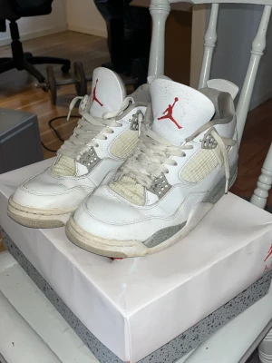 Vita Air Jordan 4 Oreo Retro sneakers - Säljer ett par klassiska Air Jordan 4 Oreo Retro sneakers i vitt läder med grå och röda detaljer. Skorna har meshpaneler på sidorna, synlig Air-sula och ikonisk Jumpman-logga på plösen. Perfekta för dig som gillar streetwear och vill sticka ut.