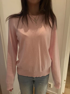 Ljusrosa stickad tröja - Mysig ljusrosa stickad tröja med rund halsringning och långa ärmar. Tröjan har ribbade muddar vid ärmslut och nederkant, vilket ger en klassisk och enkel look. Perfekt att matcha med jeans för en avslappnad stil.