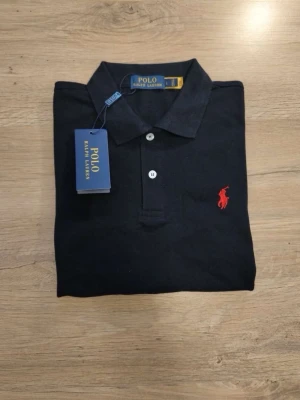 Svart pikétröja från Polo Ralph Lauren - Klassisk svart pikétröja från Polo Ralph Lauren med korta ärmar och vit knappslå. Tröjan har den ikoniska röda logotypen broderad på bröstet och är tillverkad i mjuk bomull. Perfekt till jeans eller shorts för en clean och stilren look. Det står att den är Large men den är i storleken Small 