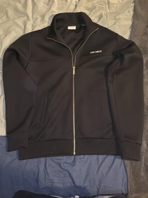 Svart jacka från Les Deux med dragkedja - Stilren svart jacka från Les Deux med hög krage och hel dragkedja framtill. Jackan har två sidofickor och diskret logga på bröstet. Tillverkad i ett mjukt material med ribbade muddar vid ärmslut och nederkant.