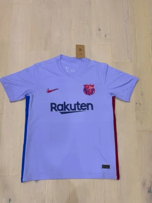 Lila FC Barcelona matchtröja Nike - Snygg lila FC Barcelona matchtröja från Nike med klubbmärke och Rakuten-tryck på bröstet. Tröjan har V-ringning, korta ärmar och detaljer i blått och rött på sidorna. Perfekt för fotbollsträning eller att bära som supporter. Tillverkad i lätt och ventilerande material.