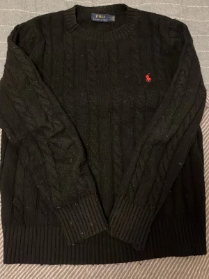 Svart kabelstickad tröja Polo Ralph Lauren - Svart kabelstickad tröja från Polo Ralph Lauren med klassisk röd broderad logga på bröstet. Tröjan har rund halsringning, långa ärmar och breda ribbade muddar vid ärmslut och nederkant. Perfekt för en stilren och tidlös look.