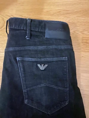 Svarta jeans från Emporio Armani - Snygga svarta jeans från Emporio Armani med klassisk femficksmodell och diskret logga på bakfickan. Jeansen har raka ben och är tillverkade i ett mjukt denimtyg som ger en skön passform. Perfekta för dig som gillar stilrena och tidlösa plagg.