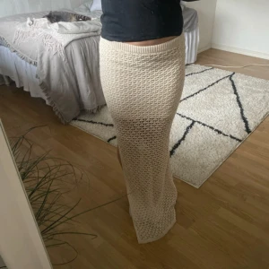 Beige stickad långkjol - Säljer en beige stickad långkjol med virkat utseende och hög midja. Kjolen har en rak passform och slits på sidan, perfekt för en trendig och avslappnad look.