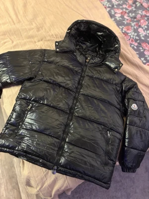 Svart dunjacka från Moncler - Snygg svart dunjacka från Moncler som aldrig är andvänd, med glansig finish, huva och dragkedja framtill. Jackan har två framfickor med dragkedja och Moncler-logga på ärmen.