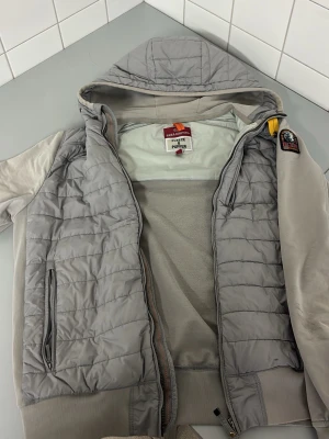 Ljusgrå quiltad jacka Parajumpers - Snygg ljusgrå quiltad jacka från Parajumpers med dragkedja framtill och en bröstficka med dragkedja. Jackan har släta ärmar och ett emblem på ena ärmen. Perfekt för dig som vill ha en stilren och bekväm jacka i vardagen. Äkta i jätte bra skick inga skador nyskick pris går DISKUTERA!!