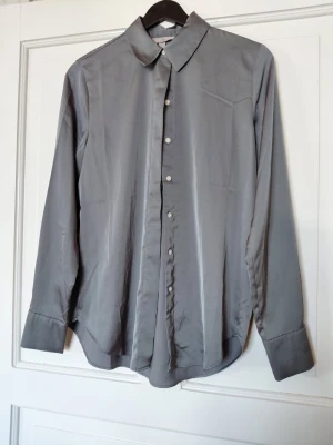 Grå silkeslen skjorta med knappar - Stilren grå skjorta i silkesliknande material med klassisk krage och långa ärmar. Skjortan har blanka knappar framtill och en mjuk, glansig finish som ger en lyxig känsla. Perfekt för dig som gillar minimalistisk och clean stil.