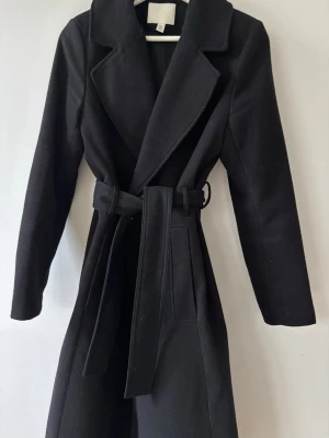 Svart lång kappa med skärp - Stilren svart lång kappa med breda slag och knytskärp i midjan. Kappan har två sidofickor och en klassisk rak passform. Perfekt för kyligare dagar och ger en snygg siluett. Materialet ser ut att vara ullblandning.