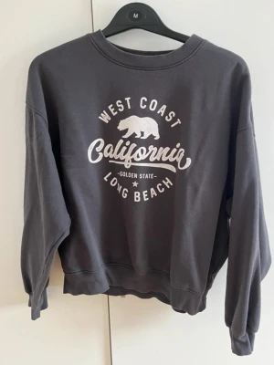 Grå sweatshirt från Bik Bok - Mysig grå sweatshirt från Bik Bok med tryck i vitt framtill: 'West Coast California Long Beach' och en björn. Tröjan har rund hals, långa ärmar och ribbade muddar. Perfekt för en avslappnad och cool stil.
