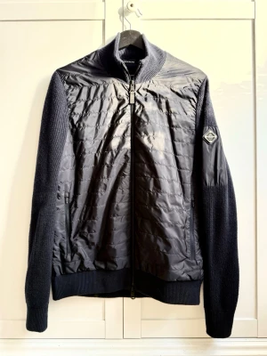 Svart hybridjacka från J.Lindeberg - Snygg svart hybridjacka från J.Lindeberg med quiltad front och stickade ärmar. dragkedja framtill och diskret logomärke på ärmen. Perfekt till vår/sommar