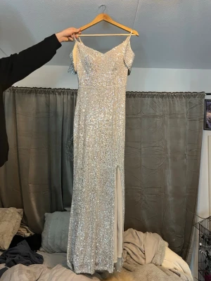 Glittrig guldklänning med slits - Snygg långklänning i guld med glittriga paljetter över hela klänningen. Offshoulder-modell med smala axelband och en elegant slits framtill. Perfekt för dig som vill sticka ut på bal eller fest. Klänningen har figurnära passform och är tillverkad i syntetmaterial. Köpte klänningen för 3000 kr till min bal