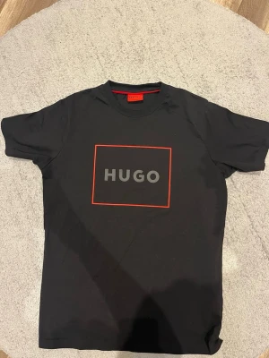 Svart HUGO t-shirt med röd ram - Snygg svart t-shirt från HUGO med en tydlig logga i vitt och grått på bröstet, inramad av en röd fyrkant. Klassisk passform och rund hals. Perfekt till jeans eller shorts för en clean och modern look.