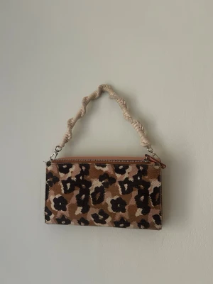 Kuvertväska med leopardmönster - Säljer en trendig kuvertväska med leopardmönster i bruna, beige och svarta toner. Väskan har en grov, flätad handrem i beige tyg och stängs med dragkedja. Perfekt accessoar för att lyfta din outfit med lite extra attityd.