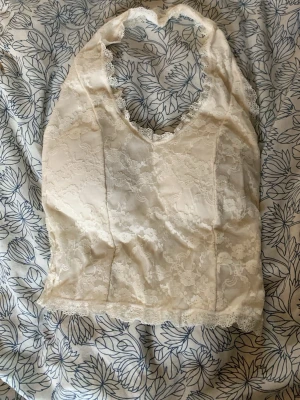 Vit spetstopp med halterneck - Säljer en vit spetstopp med halterneck. Framsidan har ett romantiskt spetsmönster och baksidan är smockad för extra komfort. Väldigt stretchig med en inbyggd bh. 