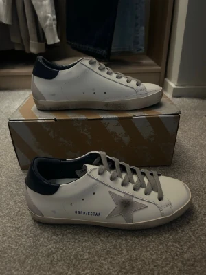Golden Goose vita sneakers med stjärna - Snygga vita sneakers från Golden Goose med grå stjärndetalj på sidan, grå skosnören och mörkblå häl. Klassisk låg modell i läder med GGDB/SSTAR-tryck på sidan och slitna detaljer för en cool look.