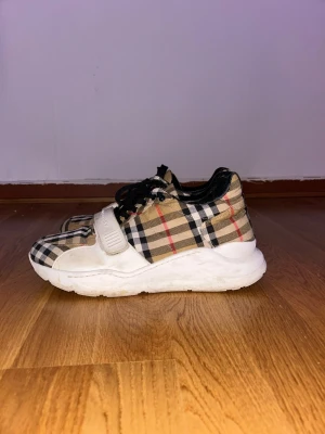 Burberry rutiga sneakers - Säljer ett par klassiska Burberry sneakers med det ikoniska beige, svarta och röda rutmönstret. Skorna har vita sulor, svarta skosnören och en vit rem med Burberry-logga över foten. Ovandelen är i tyg och detaljer i läder.