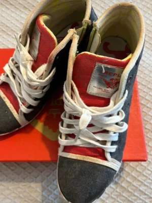 Replay sneakers i svart och rött - Snygga sneakers från Replay med svart mockaliknande material, röda detaljer och vita partier. Skorna har vita snören och en hög modell med tydlig Replay-logga på plösen. Perfekta för dig som gillar streetstyle och vill sticka ut.