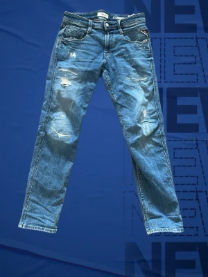 Blå slitna jeans från Replay - Säljer ett par blå jeans från Replay med slitningar och distressed detaljer på benen. Klassisk femficksmodell med ljus tvätt och snygga sömmar på bakfickan. Perfekta för en avslappnad och trendig look. Storlek W28 L30