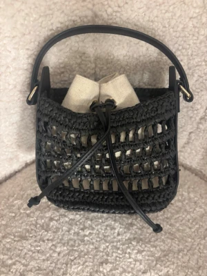 Svart flätad handväska med axelrem - Snygg svart handväska i flätad design med justerbar axelrem och kort handtag. Väskan har en beige innerpåse i tyg med dragsko. Perfekt för dig som gillar trendiga detaljer och en unik look. Passar till många olika outfits och är lätt att bära med sig.