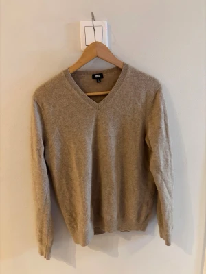 Beige v-ringad stickad tröja Uniqlo i kashmir - Mjuk och stilren beige stickad tröja från Uniqlo med v-ringning. Tröjan har långa ärmar och ribbade muddar vid ärmslut och nederkant. Materialet är i kashmir och känns lyxigt och passar bra till kyligare dagar. Tröjan är i storlek L, men passar snarare S/M.
