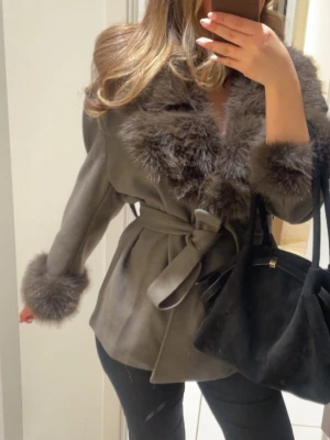 Grå kappa med fuskpäls och knytskärp - Stilren grå kappa med bred fuskpäls vid krage och ärmslut. Kappan har ett matchande knytskärp i midjan som ger en snygg siluett. Perfekt för dig som vill ha en trendig och elegant look. Pälsen är fluffig och ger extra lyxig känsla.