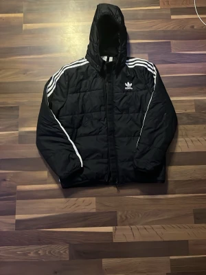 Svart Adidas vindjacka med huva - Svart vindjacka från Adidas med huva och klassiska vita ränder längs axlarna. Jackan har dragkedja framtill, quiltad design och Adidas-logga på bröstet. Perfekt för en sportig och avslappnad stil. Passar personer som är mellan 158-168 cm långa.