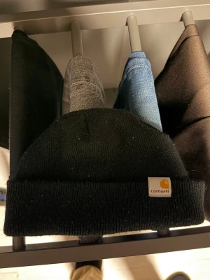 Svart mössa från Carhartt - Säljer en klassisk svart mössa från Carhartt med uppvikt kant och den ikoniska Carhartt-loggan framtill. Mössan är ribbstickad och gjord i mjukt material som sitter skönt på huvudet. Perfekt för dig som vill ha en stilren och enkel accessoar.