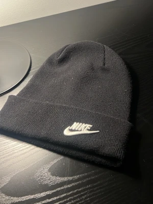 Svart mössa från Nike med broderad logga - Säljer en svart ribbstickad mössa från Nike med uppvikt kant och vit broderad Nike-logga framtill. Klassisk och enkel design som passar till det mesta. Perfekt för kalla dagar och streetwear-stil.