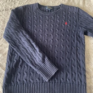 Mörkblå kabelstickad tröja Polo Ralph Lauren - Kabelstickad mörkblå tröja från Polo Ralph Lauren med rund halsringning och ribbade muddar. Klassisk röd logga broderad på bröstet. Perfekt till jeans eller kjol för en stilren look. Storlek L i barn! 14-16 år. 
