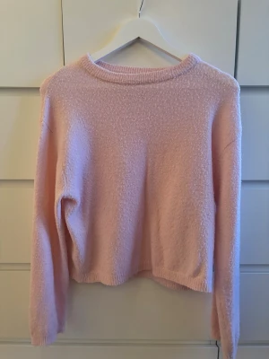 Ljusrosa fluffig stickad tröja - Mysig ljusrosa stickad tröja med rund hals och långa ärmar. Tröjan har en fluffig, mjuk struktur och ribbade muddar vid hals, ärmslut och nederkant. Perfekt för att hålla värmen under kyliga dagar.