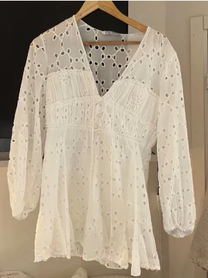 Vit broderad klänning med v-ringning - Söt vit blus med v-ringning och broderade hålmönster. Blusen har lång ärm med resår vid ärmslut och snörning framtill under bysten. Tillverkad i ett lätt och luftigt bomullstyg som ger en romantisk känsla.