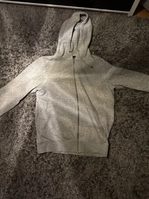 Grå zip hoodie från Jack & Jones - Snygg gråmelerad hoodie från Jack & Jones med dragkedja framtill och huva. Tillverkad i mjukt material med borstad insida för extra komfort. Perfekt för chill dagar eller när du vill ha en enkel och stilren look.