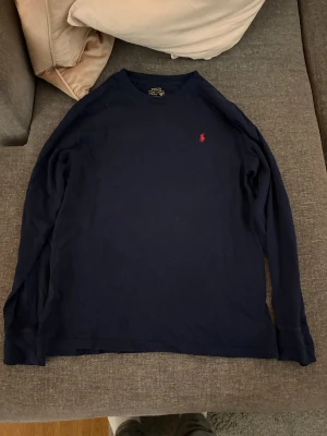 Mörkblå långärmad tröja Polo Ralph Lauren - Mörkblå långärmad tröja från Polo Ralph Lauren med klassisk röd logga broderad på bröstet. Tillverkad i mjuk bomull och har en enkel, stilren design med rund hals. Perfekt för en clean och avslappnad look.