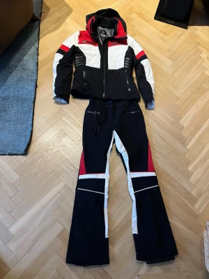 Svart, vitt och skidset från 8848 Altitude - Snyggt skidset från 8848 Altitude med jacka och byxor i svart, vitt och rött. Jackan har huva, dragkedjor och quiltade detaljer på axlarna. Byxorna har dragkedjor, fickor och vita samt röda paneler. Perfekt för dig som vill sticka ut i backen.