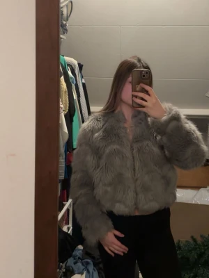 Grå kort pälsjacka - Säljer en snygg grå pälsjacka med kort modell och långa ärmar. Jackan har ett fluffigt och mjukt material som ger en lyxig känsla. Perfekt för dig som vill ha en trendig look under kalla dagar. En krok har gått av och är av någon avledning påsydd med en röd tråd, syns dock inte när man använder den, varav den låga priset