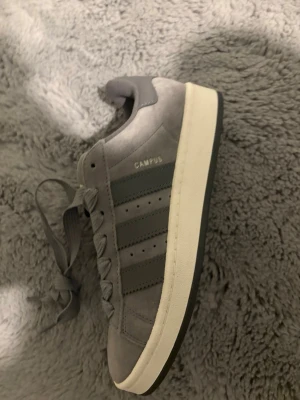 Grå Adidas Campus sneakers - Hel nya aldrig använda. Snygga grå Adidas Campus sneakers i mocka med klassiska tre ränder på sidan och vit sula. Skorna har platt tå, breda skosnören och Adidas-logga på plösen. Perfekta för en clean och stilren look.