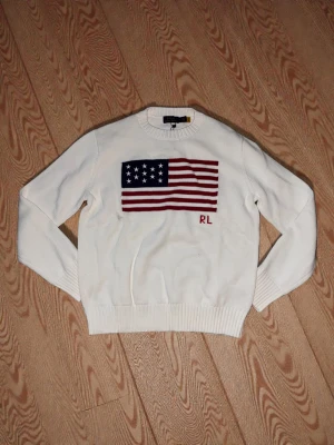 Vit stickad tröja Polo Ralph Lauren - Vit stickad tröja från Polo Ralph Lauren med amerikansk flagga i blått, rött och vitt på bröstet samt broderade initialer 'RL'. Rund halsringning och ribbade muddar. Klassisk och stilren design i mjukt material.