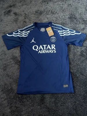 PSG matchtröja blå Jordan x Qatar - Snygg blå PSG matchtröja i samarbete med Jordan och Qatar Airways. Tröjan har vita detaljer på axlarna, klubbmärke och sponsortryck framtill samt spelarnamn och nummer 14 på ryggen. Tillverkad i lätt och funktionellt material, perfekt för fotboll eller träning.