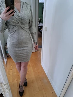 Glittrig silver kortklänning  - Säljer en glittrig silverfärgad kortklänning  och v-ringning. Klänningen har långa ärmar och markerad midja med twistad detalj framtill. Perfekt för dig som vill sticka ut på nästa event! Använd 1 gång