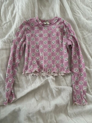 Lila ribbad långärmad topp H&M - Superfin lila ribbad långärmad topp från H&M x Smiley World med sött smiley-mönster över hela. Croppad modell med vågig kant nertill och vid ärmslut. Perfekt för dig som gillar färg och roliga prints!