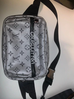 Silver axelväska från Louis Vuitton - Säljer en silverfärgad axelväska från Louis Vuitton med det klassiska monogrammönstret i svart. Väskan har en svart justerbar axelrem och en framficka med svart dragkedja där Louis Vuitton-loggan står tryckt i vitt. Materialet är glansigt och ser ut som konstläder.