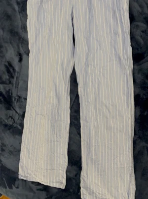 Randiga pyjamasbyxor Gina Tricot - Blå pyjamasbyxor från Gina Tricot Young med vita tunna vertikala ränder. Byxorna har resår i midjan för extra komfort och är tillverkade i ett lätt och mjukt material, perfekt för mysiga kvällar hemma.