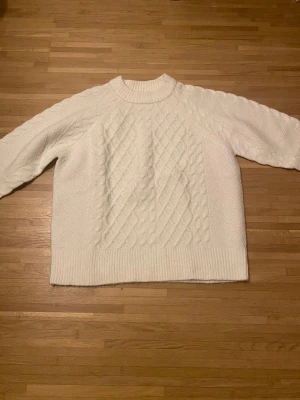 Vit kabelstickad tröja - Säljer en vit stickad tröja med kabelstickat mönster framtill och ribbade muddar vid ärmslut och nederkant. Tröjan har rund halsringning och är tillverkad i ett mjukt material som känns riktigt mysigt. Perfekt för kyliga dagar.